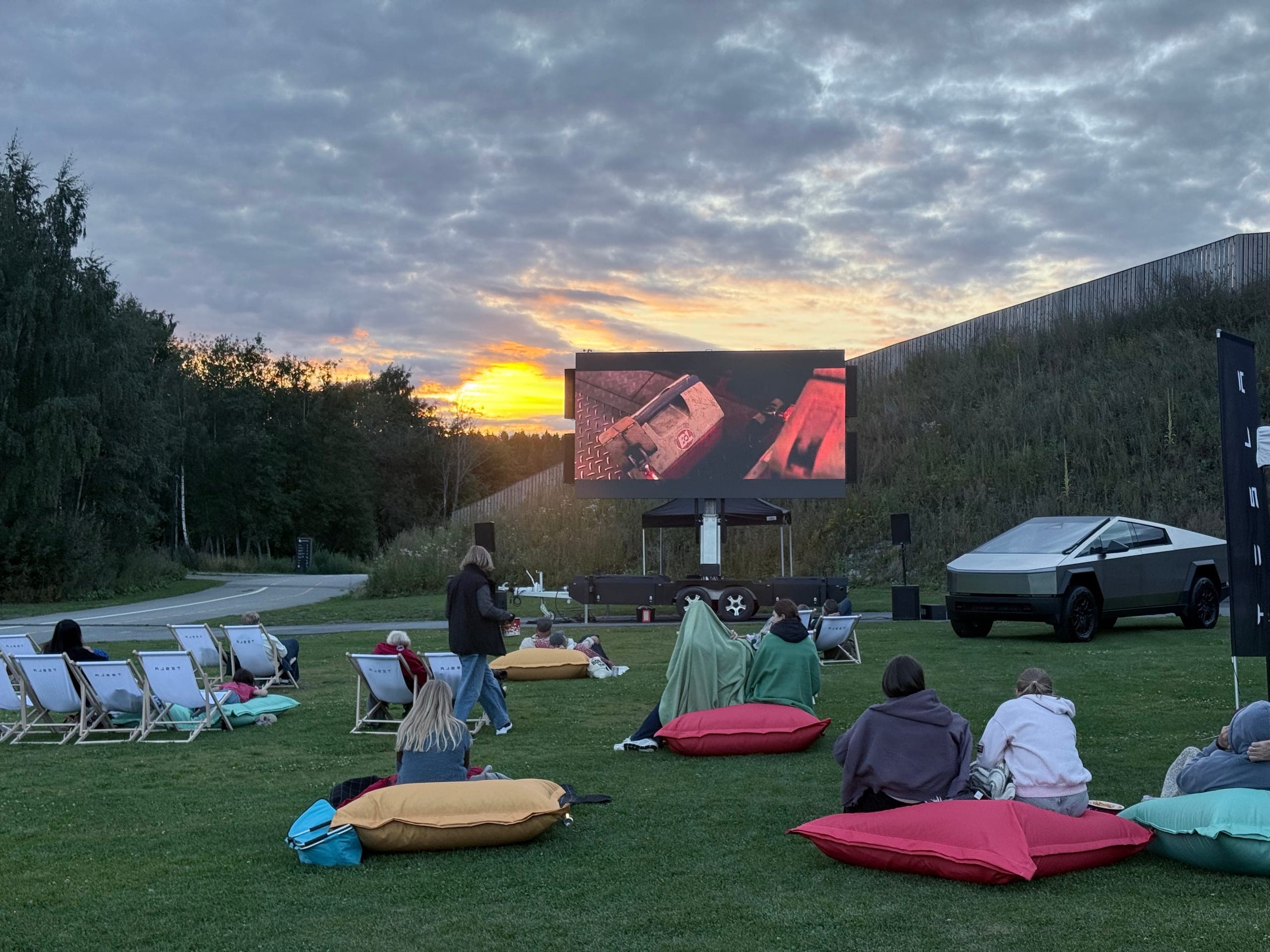 Mobil LED-skjerm på henger viser film i solnedgang mens publikum sitter på liggestoler og puter.